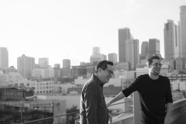 An Asian American man with short hair and glasses is laughing with a Caucasian man, with the cityscape behind them.